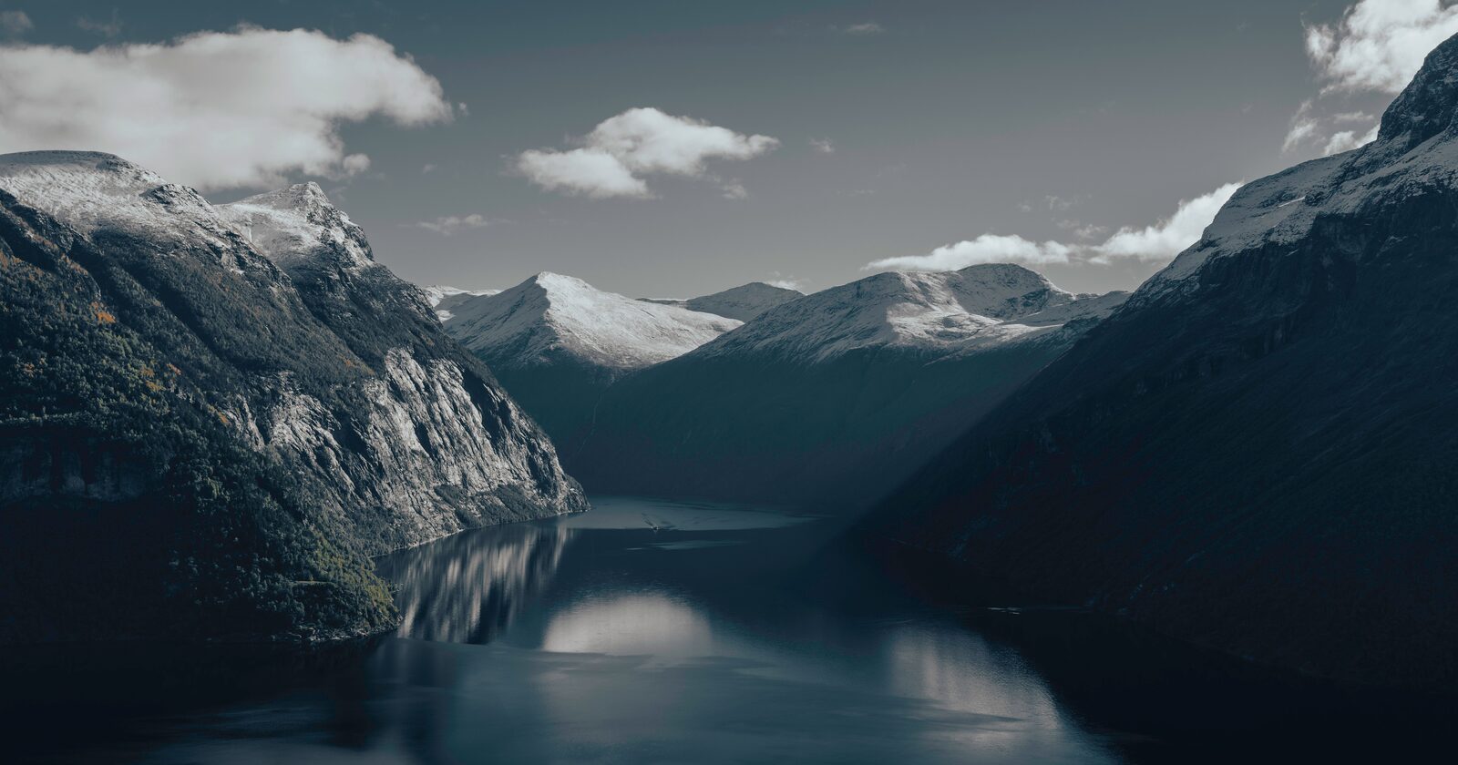 A calm Norwegian fjord flanked by snow-capped mountains under a moody sky
